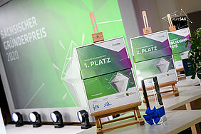 Die Urkunden und Pokale der futureSax Innovationskonferenz 2020. Im Hintergrund ist die Bühne zu sehen.
