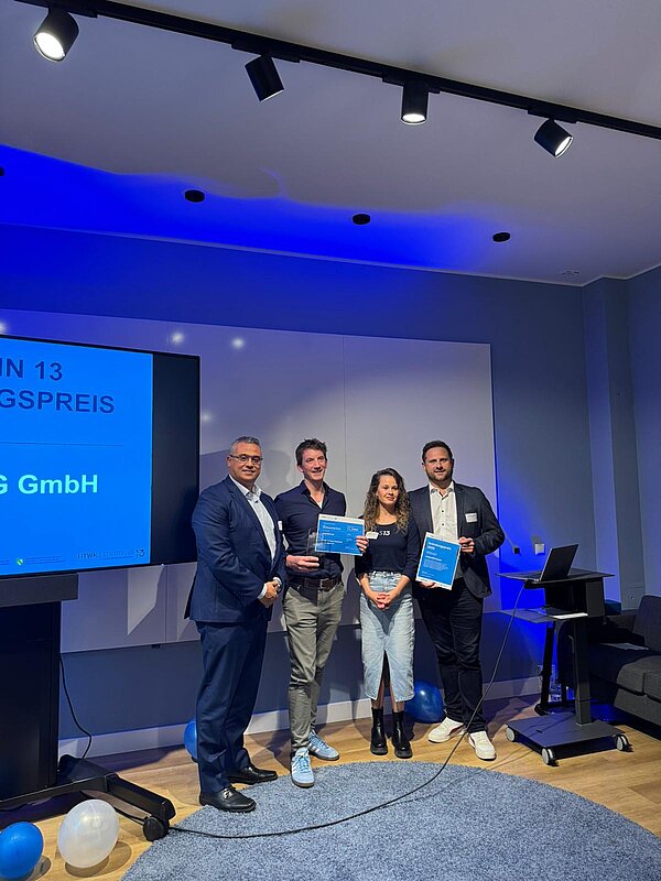 Vergabe des ersten Startbahn 13 Gründungspreises. Der Preisträger, Florian Stegmann von der AdaptING GmbH, hält Pokal und Urkunde. Neben ihm stehen Vanessa von Startbahn 13, Manuel von der Vollack Gruppe und Prof. Derbel